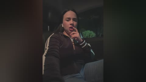 Media: Video of a young woman with long dark hair, wearing a black zip-up jacket, sitting in a dimly lit car, covering her mouth, looking distressed.