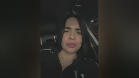 Media: Video of a woman with long black hair and medium skin tone, wearing a black jacket, sitting in a car, making a kissing face.