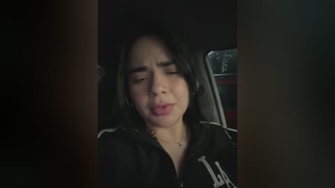 Media: Video of a young woman with medium skin tone and shoulder-length black hair, wearing a black hoodie, seated in a dimly lit car. She has a distressed expression, eyes closed, mouth slightly open.