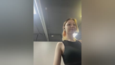 Media: Video of a fair-skinned, slender woman with shoulder-length blonde hair, wearing a black sleeveless top, standing in a dimly lit, modern room with exposed pipes and muted colors.