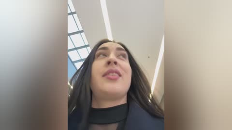 Media: A video of a woman with long, dark hair and fair skin, wearing a black blouse, taken from a low angle, showing her slightly tilted head and pursed lips. Background features a modern interior with a grid ceiling and light fixtures.