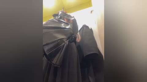 Media: A video of a person dressed in a black, shiny, raincoat with a large, black umbrella, standing in a dimly lit hallway with a yellow ceiling and white walls.