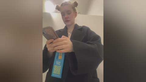 Media: A video of a woman with light skin and blonde hair styled in buns, wearing a dark blazer and holding a smartphone, inside a dimly lit room with blurred background.