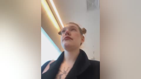 Media: A video of a young, fair-skinned woman with blonde hair in two buns, wearing a black coat, standing in a brightly lit, minimalist hallway.