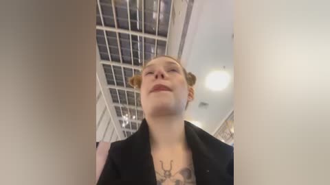 Media: Video of a smiling young woman with light skin and blonde hair styled in two small buns, wearing a black jacket, standing in an indoor space with a glass ceiling and white walls.