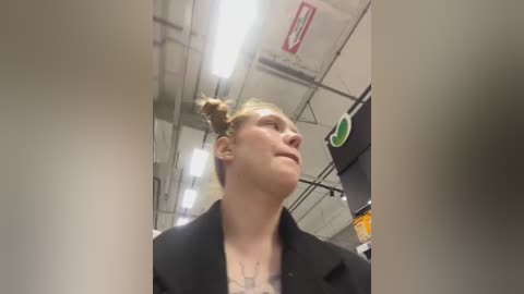 Media: A video of a woman with blonde hair in a bun, wearing a black blazer over a white shirt, looking upward in an industrial setting with exposed pipes and fluorescent lights.