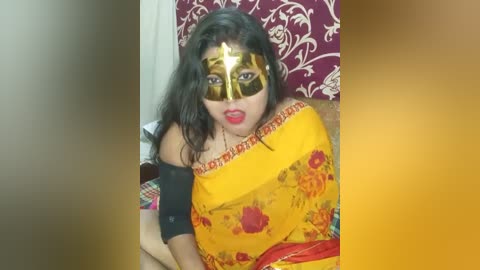 Media: Video of a South Asian woman with long dark hair, wearing a yellow sari with red floral designs, a black off-shoulder blouse, and a gold masquerade mask with red lipstick, seated against a red and white patterned backdrop.