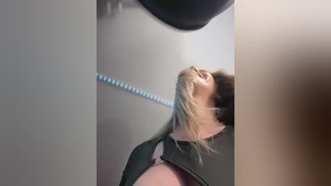 Media: Video of a woman in a black harness, her head thrown back in pleasure, against a beige wall with a blue rope hanging. Her blonde hair is tousled.