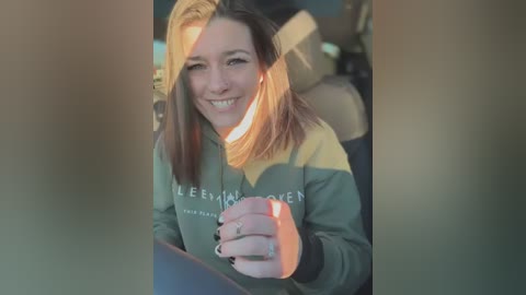 Media: Video of a smiling, light-skinned woman with shoulder-length brown hair, wearing a grey and yellow hoodie, holding a phone, inside a car.