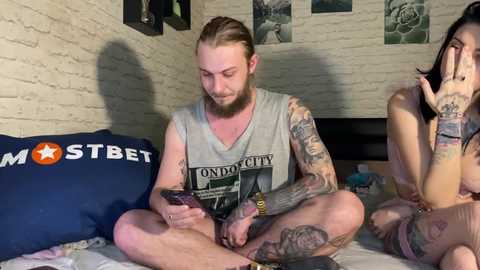 Media: Video of a tattooed man with long hair, wearing a grey tank top, sitting on a bed with a blue pillow reading \"MOSTBET.\" A partially nude tattooed woman is behind him.