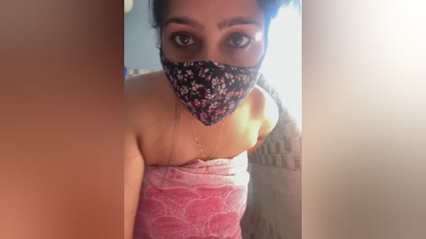 Media: Video of a woman with medium brown skin, wearing a floral face mask and a pink lace top, standing in a bright, blurred bathroom.