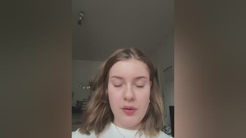 Media: Video of a young Caucasian woman with fair skin and shoulder-length brown hair, wearing a white top, closing her eyes and looking serene. Background features a dimly lit room with beige walls and indistinct furniture.