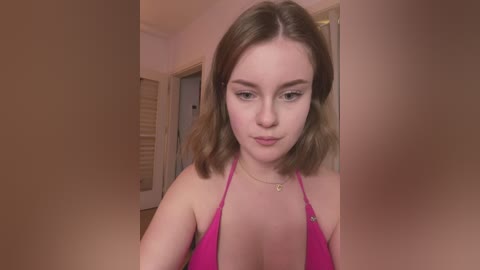 Media: Video of a young Caucasian woman with shoulder-length brown hair, wearing a pink halter top, standing in a beige-walled room with closed wooden doors and blinds.