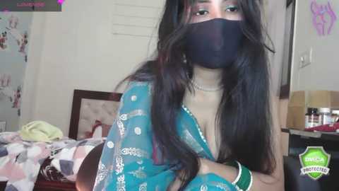 Media: A video of a South Asian woman with long black hair and fair skin, wearing a blue sari, black mask, and pearl necklace, seated in a cluttered bedroom with a bed and a desk in the background.