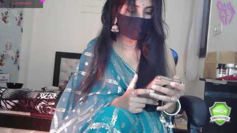 Media: Video of a South Asian woman with long black hair, wearing a turquoise saree, black face mask, and silver earrings, holding a phone. Background shows a cluttered room with a bed, posters, and a mirror.