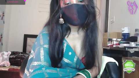 Media: Video of a woman with long black hair, wearing a teal saree with silver embroidery, a black face mask, and a choker, seated at a cluttered desk in a dimly lit room.