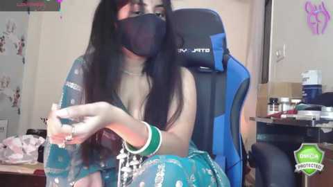 Media: Video of a young woman with long black hair, wearing a face mask, blue dress, and green wristband, playing a video game in a cluttered, brightly lit room.