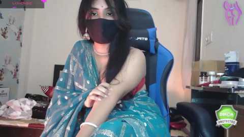 Media: A video of a woman wearing a blue sari and black mask, sitting in a gaming chair, surrounded by cluttered office supplies and posters in a dimly lit room.