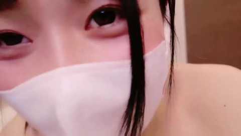 Media: Close-up video of an Asian woman with long black hair, wearing a white face mask, showing her large, expressive brown eyes. The background is blurred, suggesting an indoor setting.