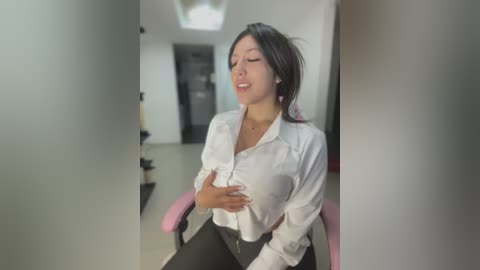 Media: Video of an Asian woman in a white blouse, sitting on a pink chair, appearing distressed, with a hand on her chest, in a modern hallway with white walls and a grey door.