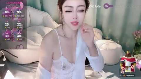 Media: Video of a fair-skinned woman with brown hair, wearing a white lace bralette and sheer robe, lying on a bed with white pillows and a green curtain background, displaying a live stream overlay.