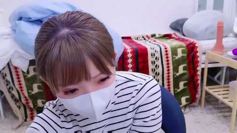 Media: Video of a young Asian woman with brown hair, wearing a white surgical mask, sitting on a bed in a simple bedroom with a green and red patterned blanket, a wooden nightstand with a sex toy, and a blue towel.