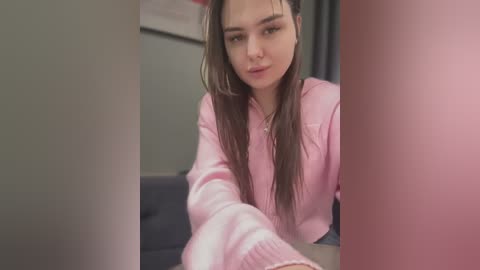 Media: A video of a young Asian woman with long brown hair, wearing a light pink hoodie, sitting on a gray couch in a dimly lit room with muted walls.