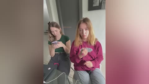 Media: Video of two teenage girls, one with blonde hair, seated indoors, engrossed in smartphones. One wears a green crop top, the other a maroon sweater.