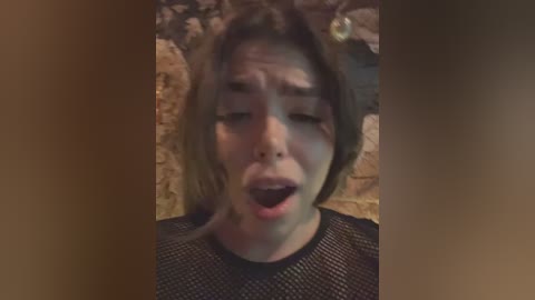 Media: Video of a Caucasian woman with light skin, short brown hair, wearing a black mesh top, in an emotional, open-mouthed expression, against a rustic stone wall background.