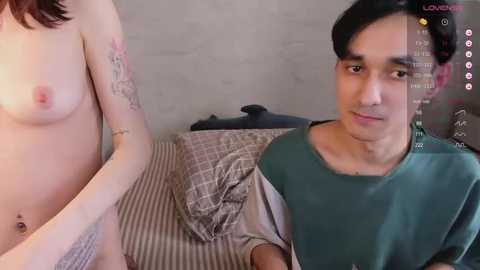 Media: Video of a nude woman with small breasts and tattoos on her arm, sitting beside a man wearing a green shirt in a dimly-lit room.