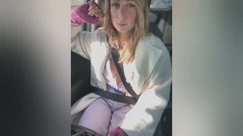 Media: Video of a blonde woman in a car, wearing a light sweater, purple gloves, and a pink scarf, seated with a concerned expression, looking to the right.