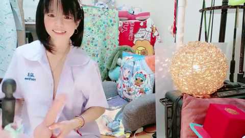 Media: Video of a smiling Asian woman with shoulder-length black hair, wearing a white polo shirt, standing in a cluttered room filled with presents, including a large, intricate paper lantern.