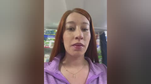 Media: Video of a young Asian woman with straight red hair, fair skin, and a slight smile. She's wearing a purple hoodie, and there's a blurred supermarket aisle behind her.