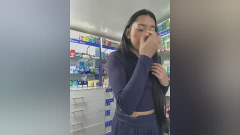 Media: Video of an Asian woman in a drugstore, covering her mouth with one hand, wearing a dark blue long-sleeve top and pants. Shelves with various products and signs in the background.