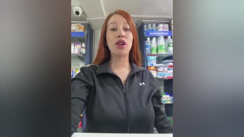 Media: Video of a young Asian woman with long red hair, wearing a black Under Armour zip-up jacket, standing behind a pharmacy counter. Shelves behind her are stocked with various products.