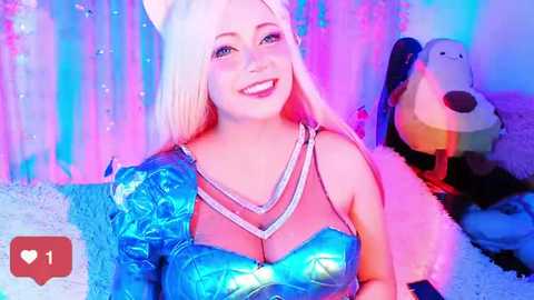 Media: A vibrant video of a smiling, light-skinned woman with long, platinum blonde hair in a glittery, blue metallic dress, posing against a backdrop of pastel-colored curtains and plush toys.