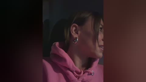 Media: Video of a young woman with light skin, brown hair tied back, smiling, wearing a pink hoodie, and dark earrings, in a dimly lit room.