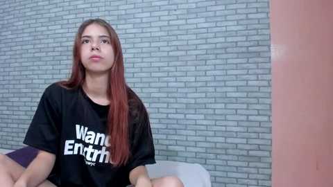 Media: Video of a young woman with long, straight auburn hair, wearing a black \"Wanna Eternity\" t-shirt, sitting against a grey brick wall, with a white chair and pink wall to the right.