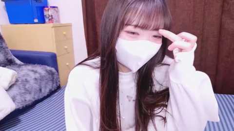 Media: Video of a young Asian woman with long dark hair, wearing a white mask, long-sleeved white top, and sitting on a blue and white striped bed in a bedroom with brown curtains and a beige dresser.