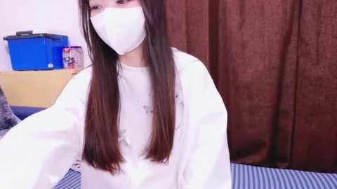 Media: Video of a young Asian woman with long black hair, wearing a white surgical mask and gown, seated on a bed with blue and white striped sheets. Background includes a blue box and brown curtains.