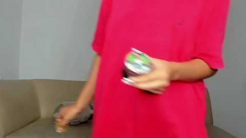 Media: Video of a person in a bright pink dress, holding a can of beer. The background features a beige couch and white walls. The individual has manicured nails and is mid-movement.