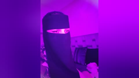 Media: Video of a person wearing a black burqa in a room with purple lighting, partially obscuring their face. Background features a bed with a white headboard and scattered pillows.