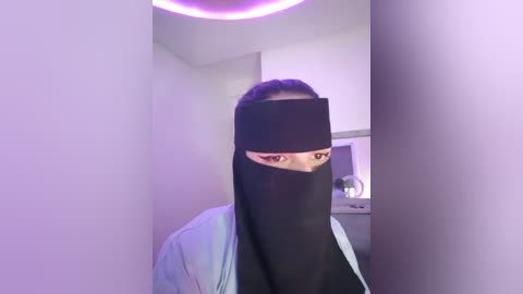 Media: Video of a woman with long dark hair and brown eyes, partially covered by a black niqab, wearing a light blue jacket. She stands in a minimalist, white-walled room with purple lighting.