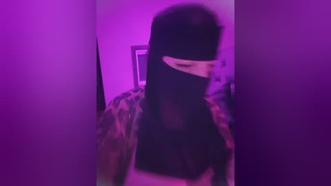 Media: A blurry, low-resolution video of a person wearing a black face mask and camouflage jacket, bathed in purple light, standing in a dimly lit room.