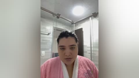 Media: A video of a middle-aged person with a neutral expression, wearing a pink bathrobe, standing in a modern, tiled bathroom with a glass shower and white walls.