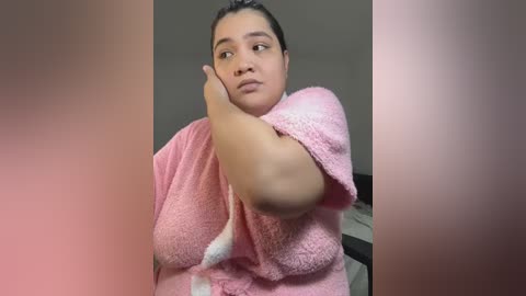 Media: Video of a plus-size woman with medium brown skin, wearing a pink fluffy bathrobe, drying her dark hair. She has a contemplative expression. Background is blurred.