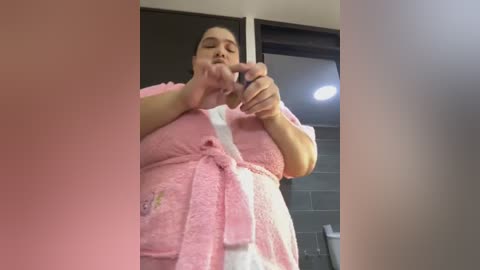 Media: Video of a plus-sized woman with fair skin, dark hair, wearing a pink bathrobe, holding a small object in her mouth, standing in a modern bathroom with tiled walls and a sink.