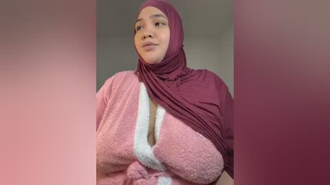 Media: Video of a light-skinned woman with a medium build, wearing a maroon hijab and a pink, fluffy bathrobe. She has a calm expression, and the background is a gradient of soft pink and beige tones.