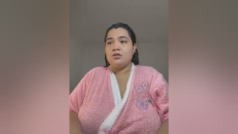 Media: Video of a plus-sized woman with dark hair, wearing a pink, fuzzy bathrobe with butterfly embroidery, standing in a neutral, blurred background.