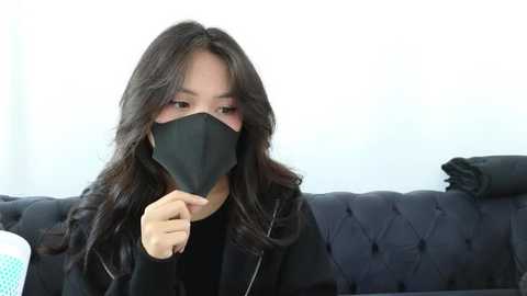 Media: Video of an Asian woman with long black hair wearing a black face mask, black jacket, and black top, sitting on a black tufted sofa, with a white wall in the background.